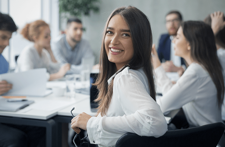 A woman in a white shirt sits at a meeting table, with colleagues discussing around her in a modern office setting.
