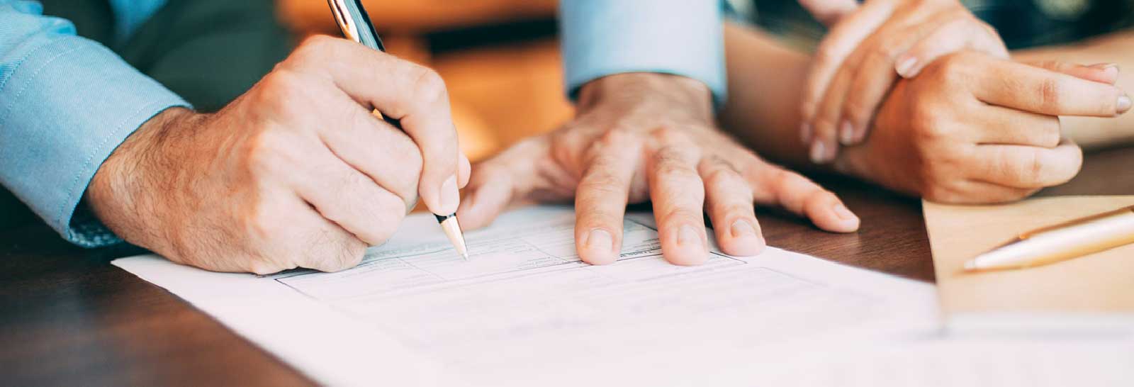 A hand writes on a contract with a pen while another hand rests on the paper. A notebook and a pen are visible in the background.