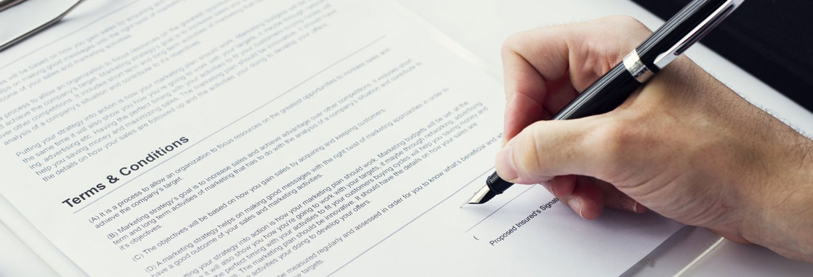 Dłoń trzymająca pióro podpisuje dokument zatytułowany 'Terms & Conditions', z widocznym szczegółowym tekstem na stronie.