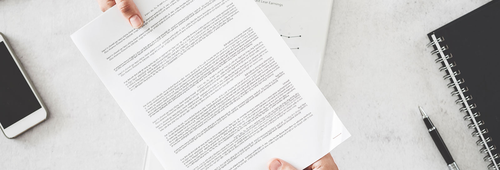 Osoba trzyma biały dokument z tekstem, podczas gdy na biurku widoczny jest smartfon, notes i długopis.