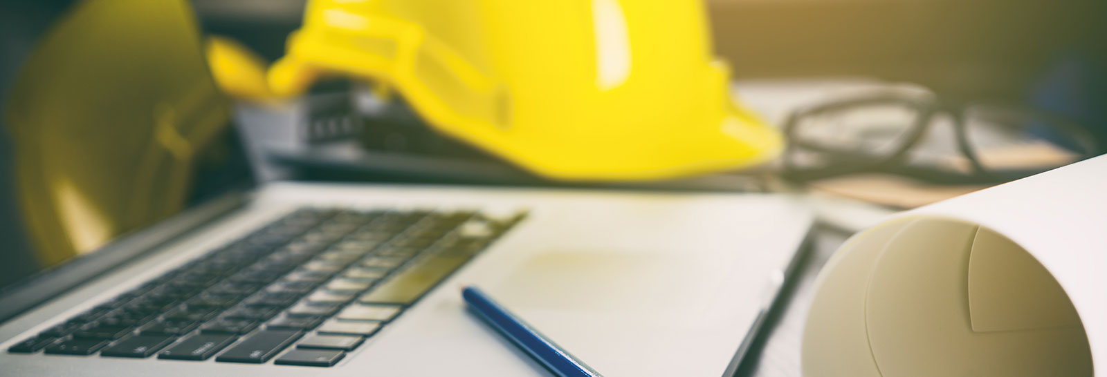 A close-up of a laptop keyboard with a yellow hard hat, a rolled-up document, and a blue pen on a work desk.