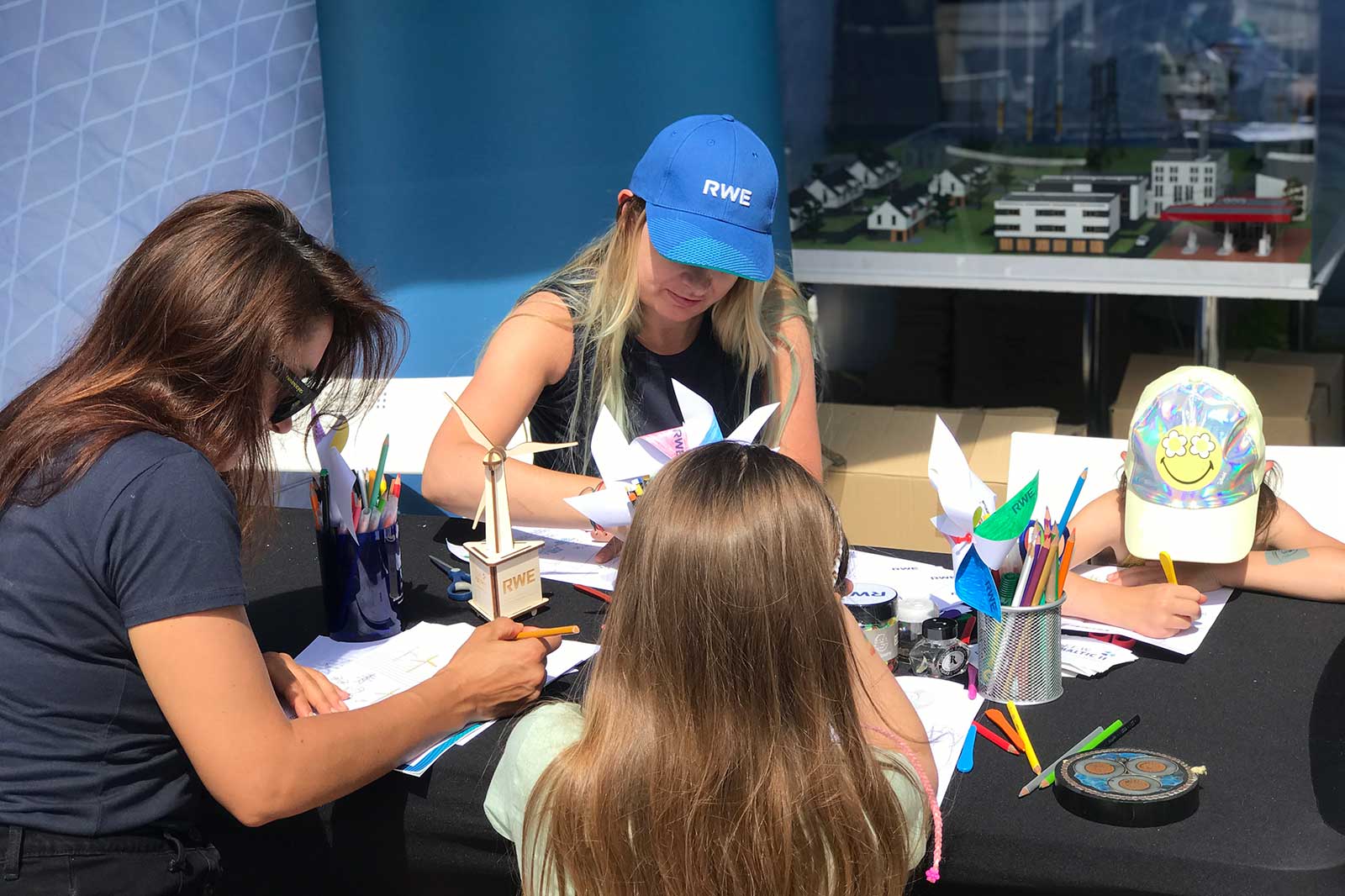 Three children and an adult are engaged in arts and crafts at a table filled with colourful pens and papers, featuring a model display in the background.