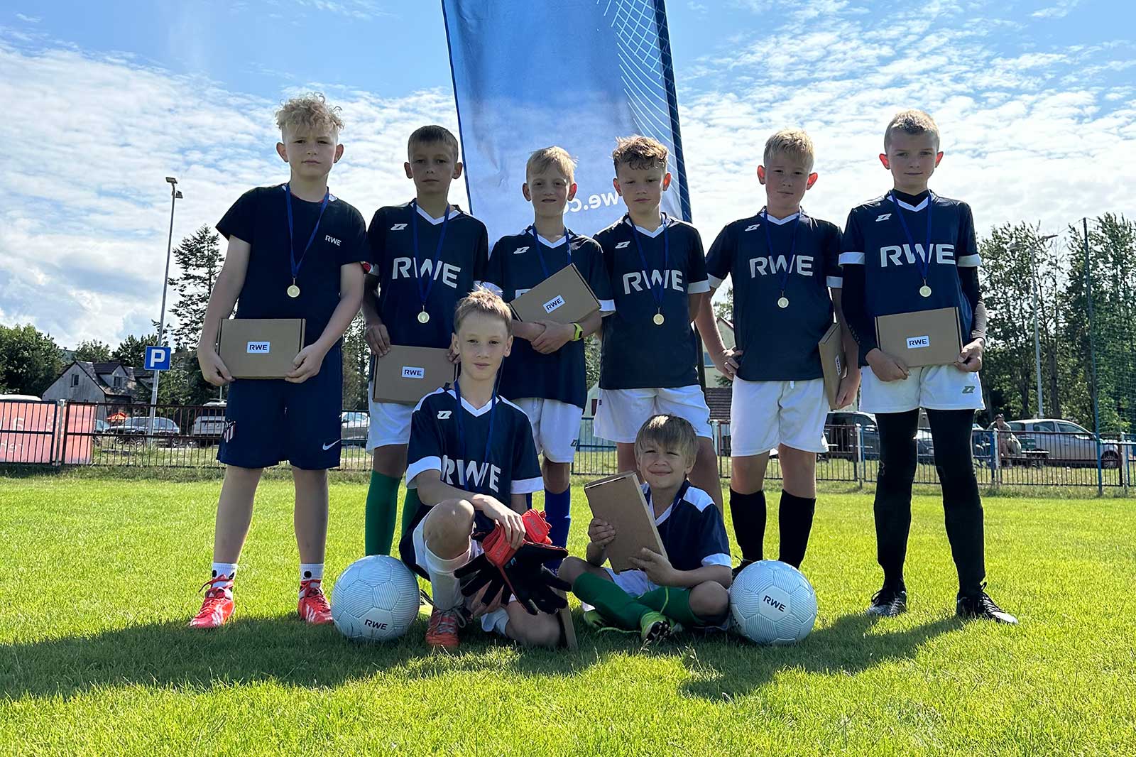 A group of children in football kits holding certificates, standing on grass with footballs and a RWE banner in the background.