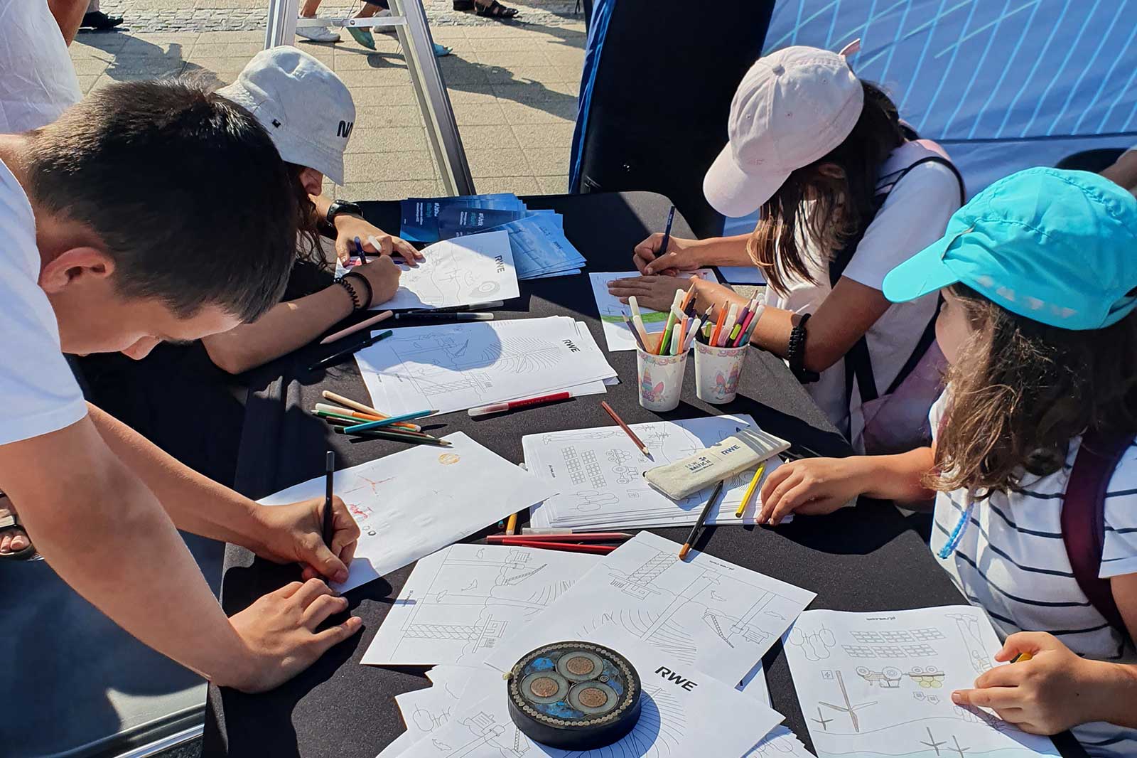 Children with different hats draw on sheets of paper at a table, surrounded by coloured pencils. Some papers have the word RWE.