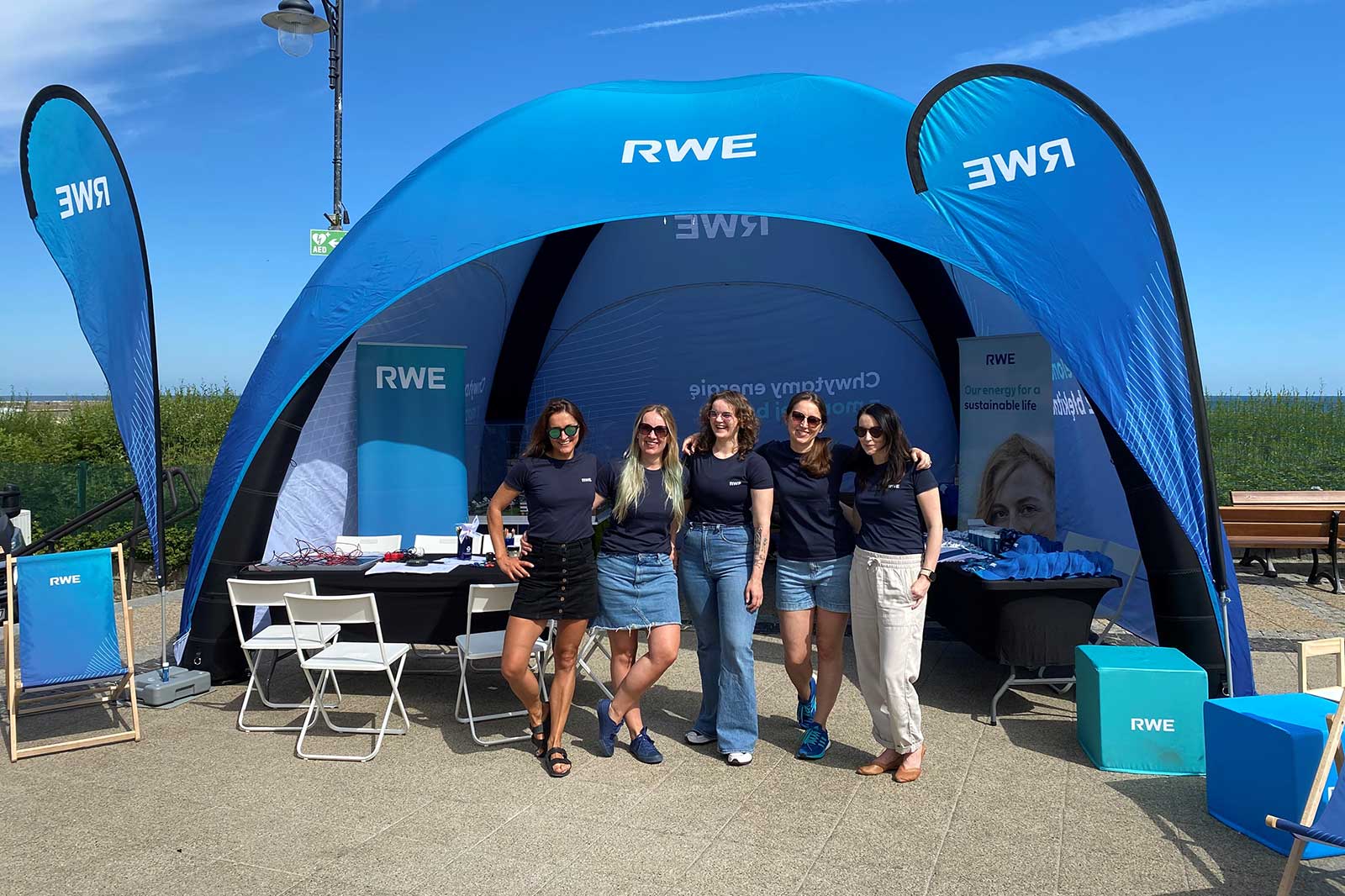 A group of five women in blue RWE shirts stand in front of a blue RWE tent with promotional materials and seating.
