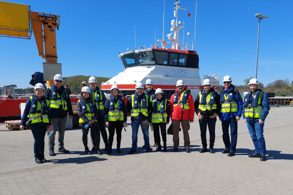 A group of people in safety gear stands in front of a boat at a port under clear blue skies.