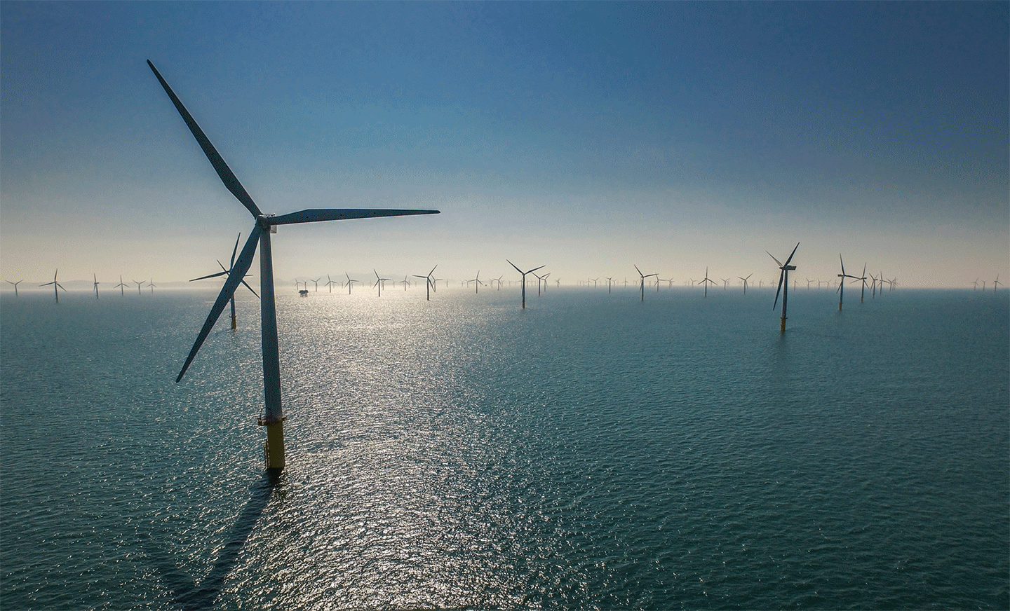 Aerial view of offshore wind turbines standing in calm waters under a clear blue sky, with reflections on the water surface.
