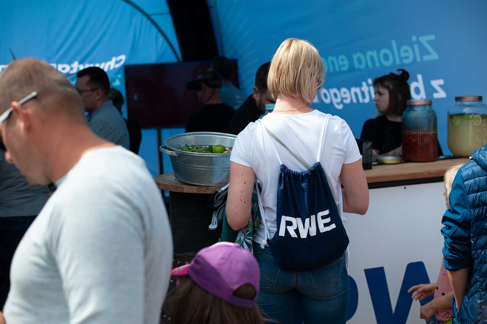 A woman wearing a white shirt and a blue RWE backpack stands in front of a food stall serving beverages and snacks.
