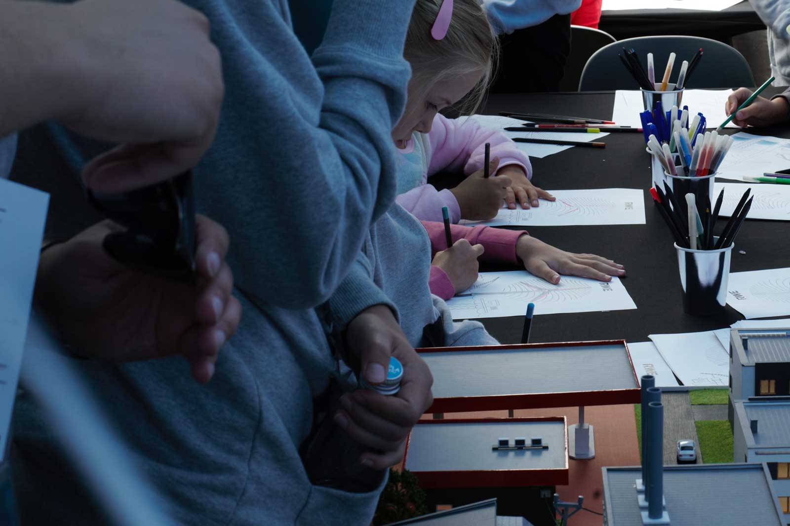 A table with children drawing and writing. Various coloured pens are placed in cups on the table.