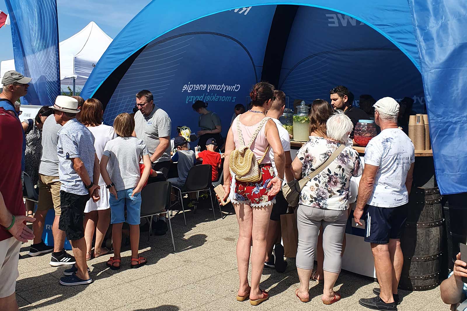 A crowd gathers at a vibrant tent, engaging with staff and enjoying refreshments in a sunny outdoor setting.