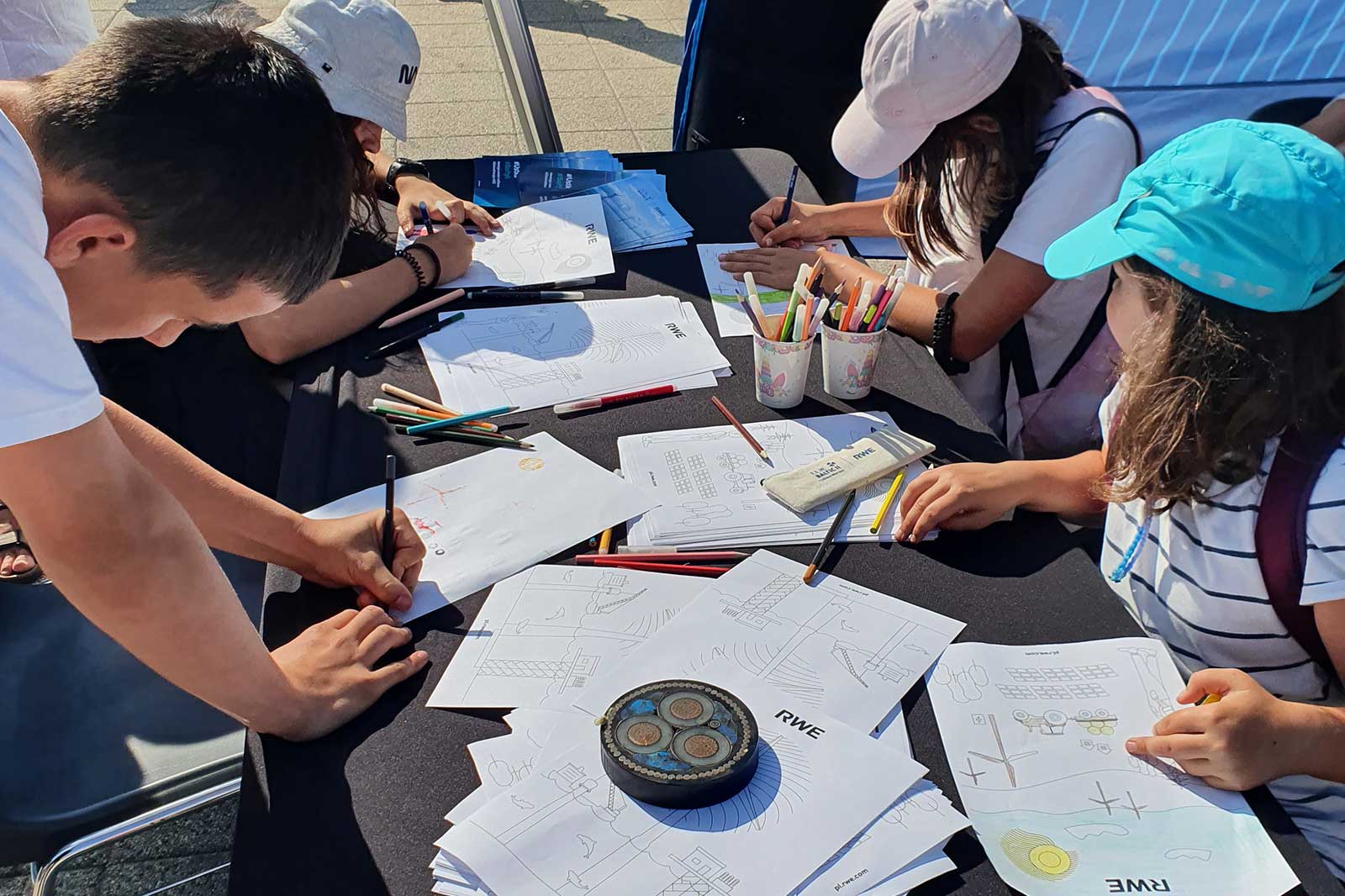 Children are drawing on paper with coloured pencils at a table. There are cups of pencils, and sheets featuring 'RWE'.