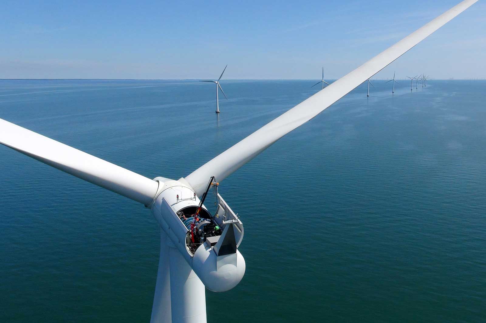 Aerial view of a wind turbine with blades, overlooking calm water and multiple wind turbines in the distance under a clear blue sky.