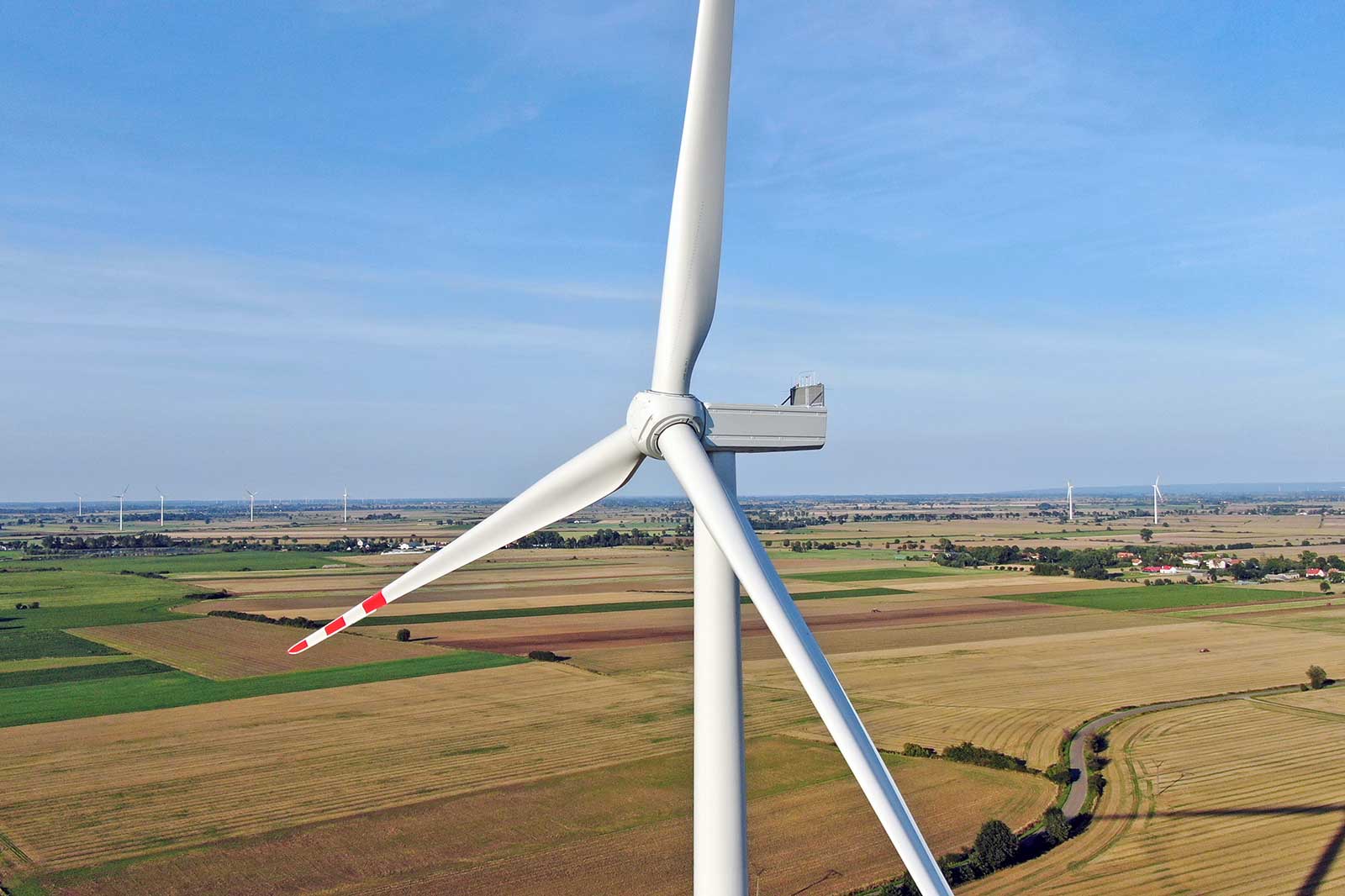 A close-up of a wind turbine in a green and brown agricultural landscape with fields and distant wind turbines under a blue sky.