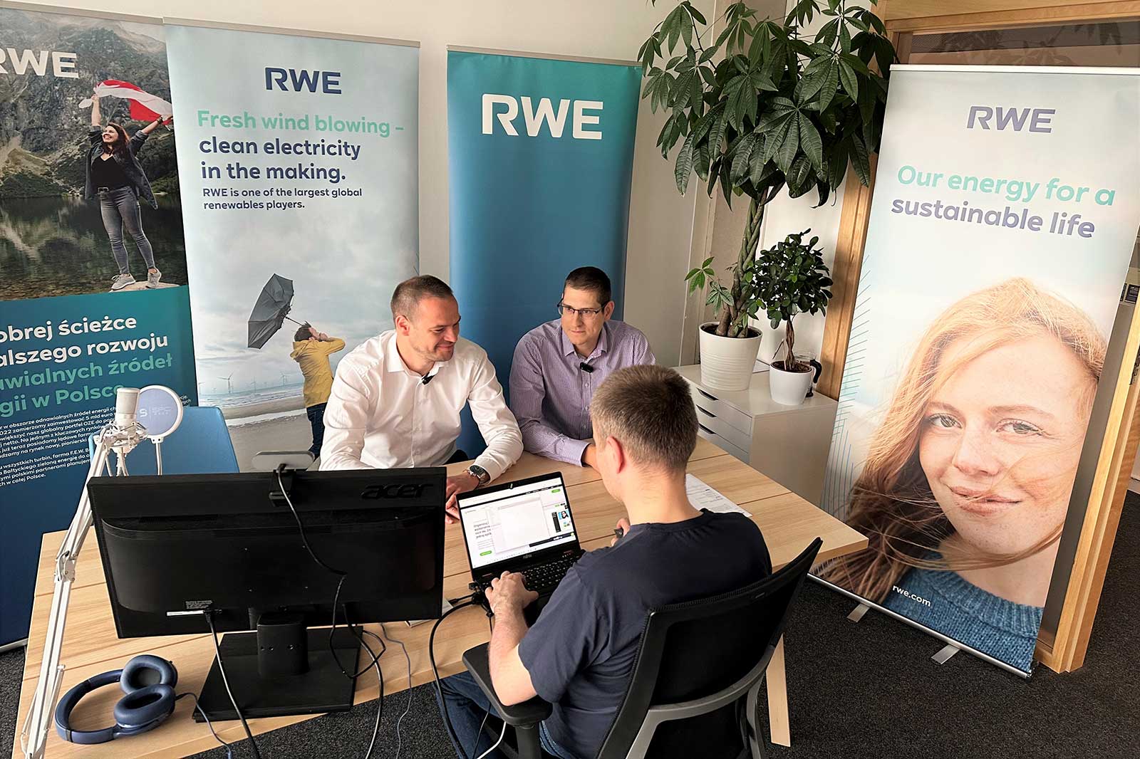 Three people engaged in a discussion in an office setting, with RWE banners promoting clean electricity and sustainability in the background.