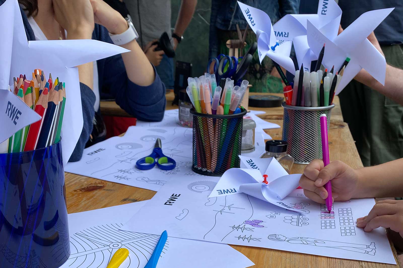 A creative setup with colourful pencils, pens, and paper illustrations. A child draws while paper pinwheels are on display.