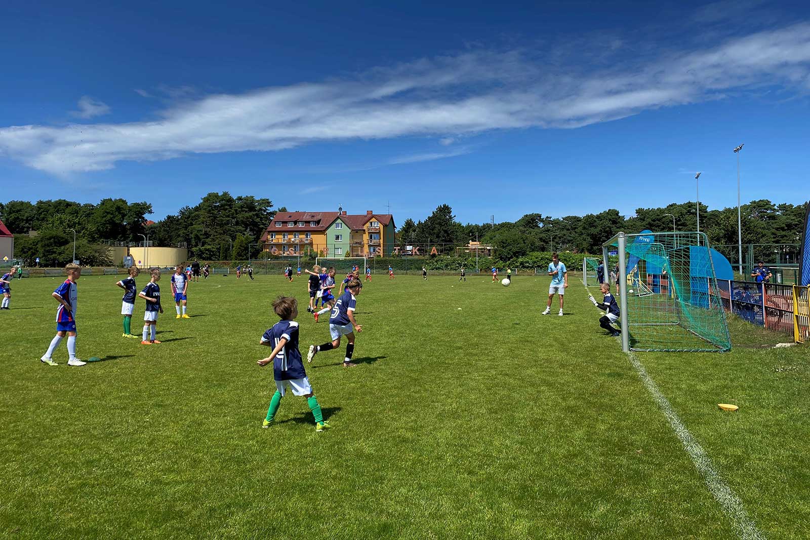 Children play football on a green field under a blue sky. A coach instructs a goalkeeper near a goal.