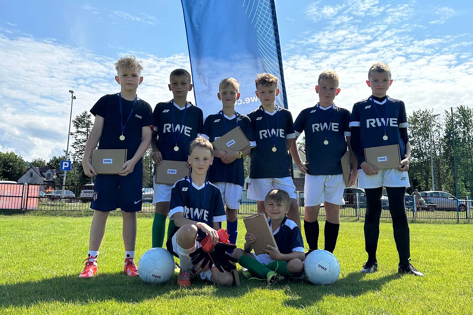 A group of children in football kits holding certificates and medals while standing on a grassy field. A banner reads 'we can'.