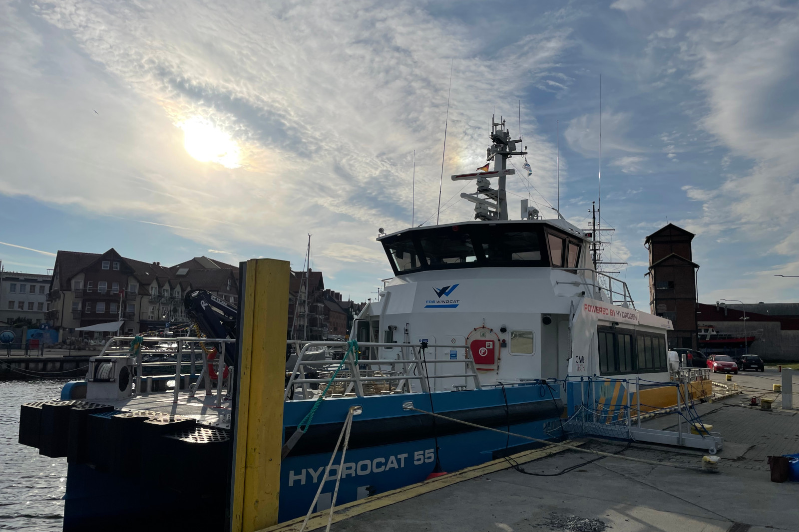 A modern hydrogen-powered boat named Hydrocat 55 docked at a harbour with buildings and a waterway in the background.