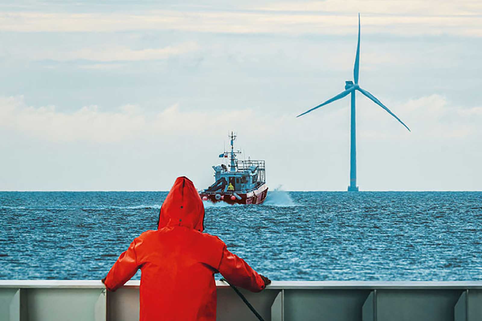 Osoba w czerwonej kurtce obserwuje nadpływający statek w kierunku dużej turbiny wiatrowej na morzu pod pochmurnym niebem.