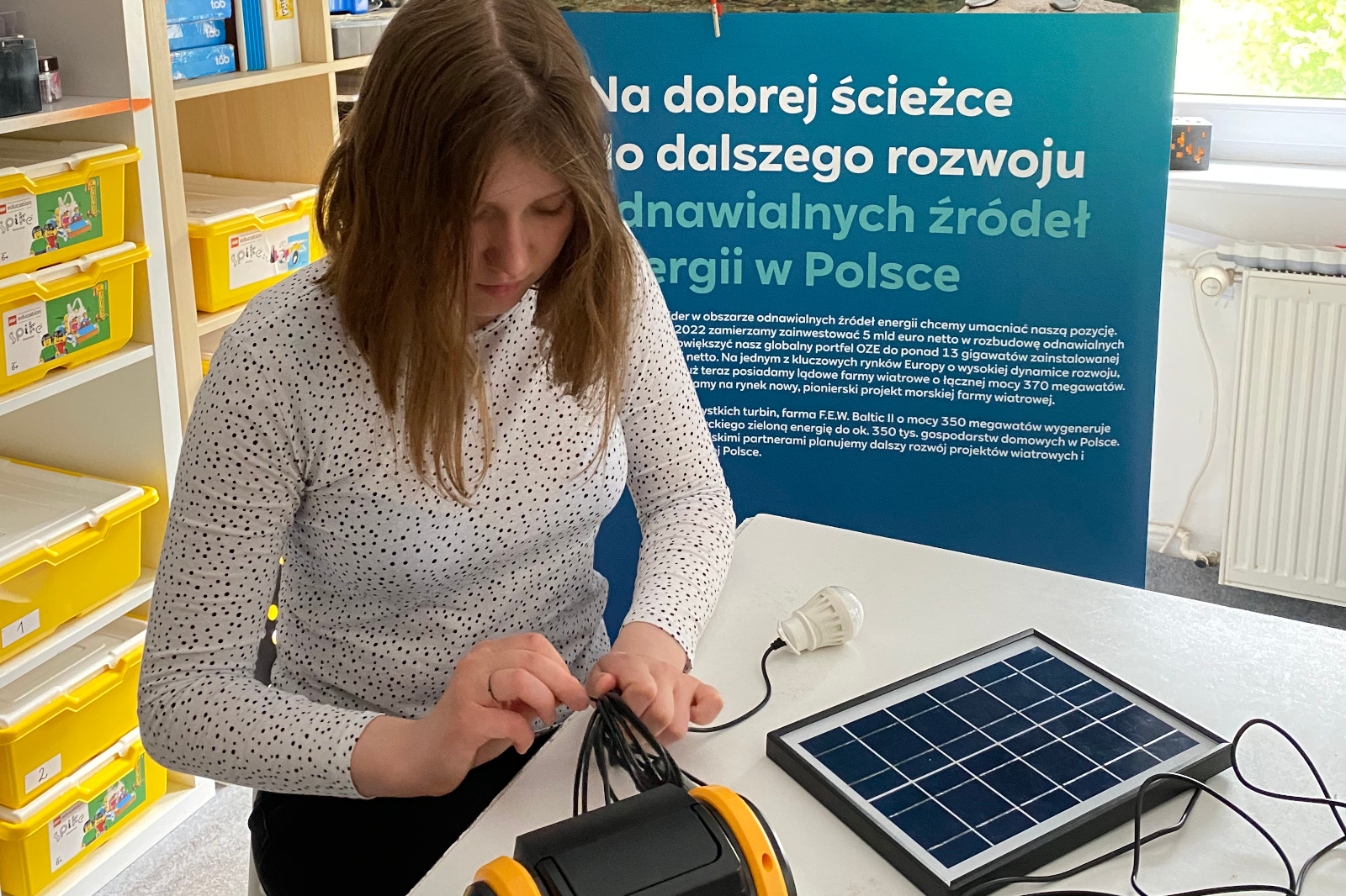 A person connects cables to a solar panel and a device on a table, with educational materials and a green energy banner in the background.