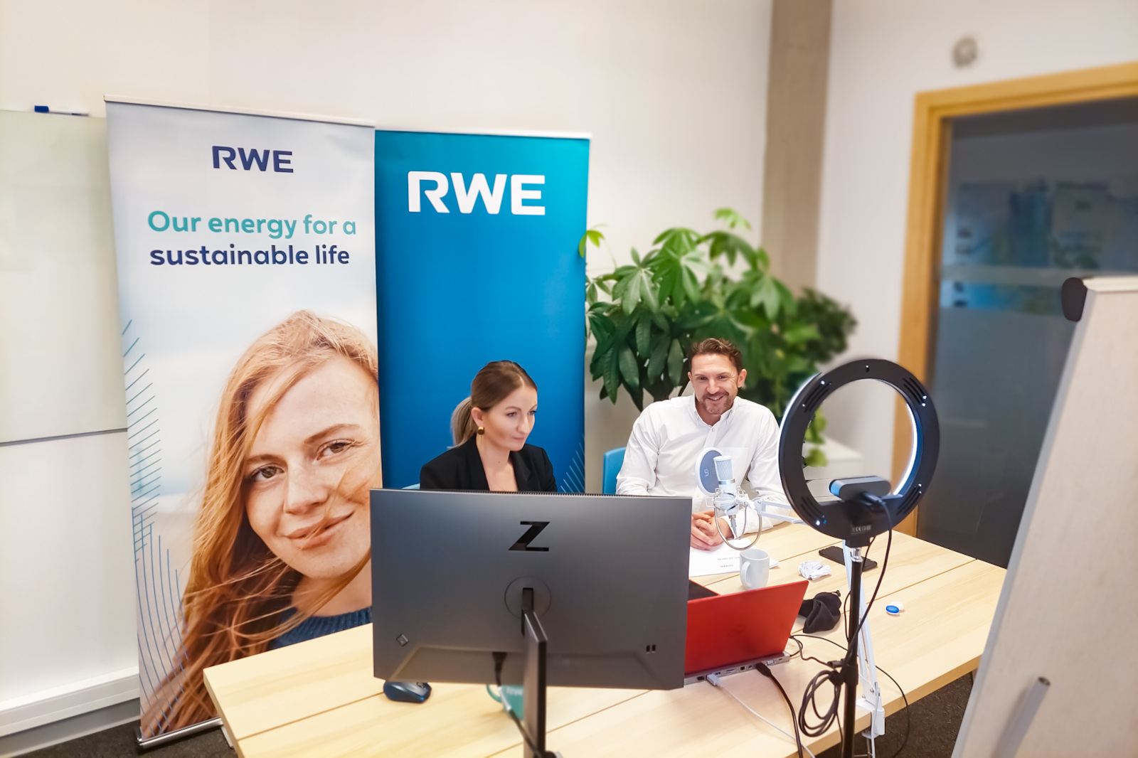 A conference setting with RWE branding, featuring two individuals at a desk with a computer, microphone, and plant backdrop.