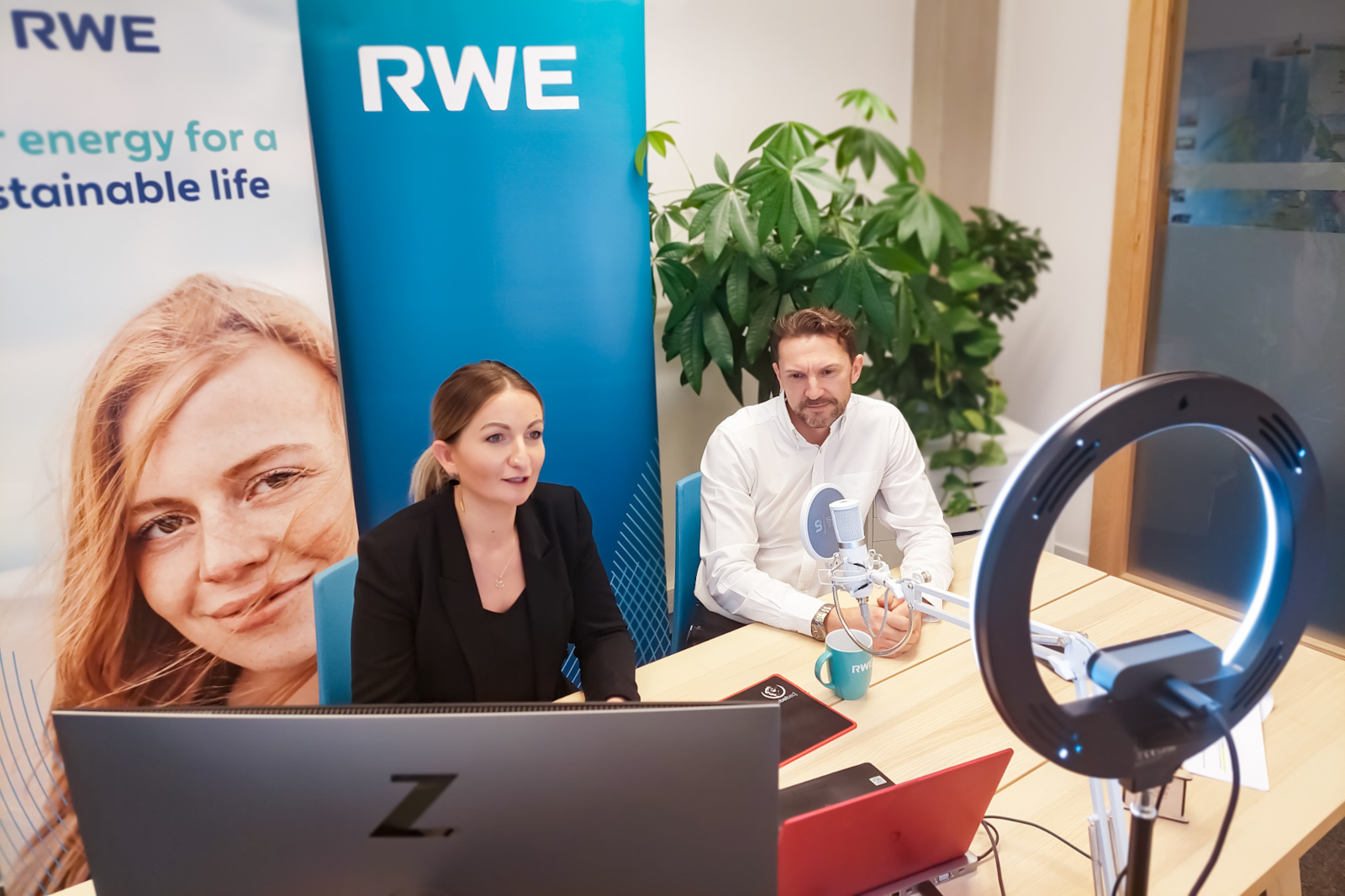 Two professionals sit at a desk with a computer, a microphone, and a ring light, surrounded by plants and an RWE banner.