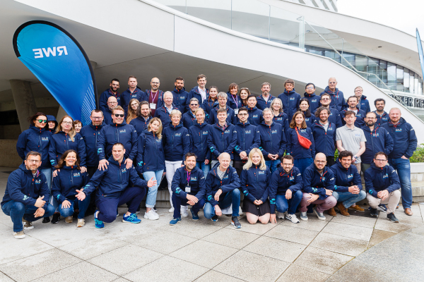 A large group of smiling individuals in matching navy jackets pose together outdoors near a building with a blue flag in the background.