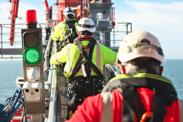 Three workers in safety gear walk along a platform with a green traffic light in the foreground, overlooking the ocean.